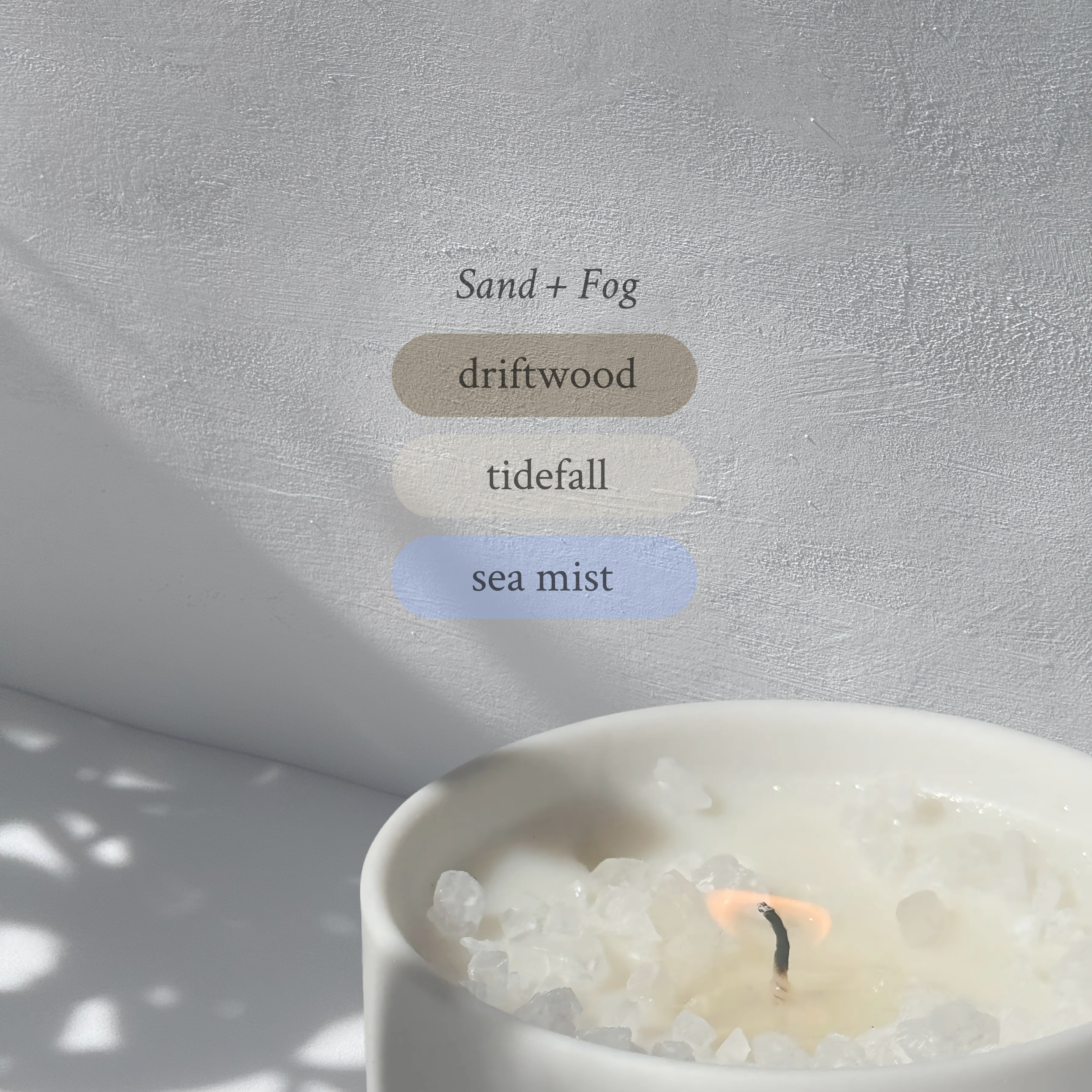 Large stoneware candle, lit, it’s top covered in rock salt. The title reads “Sand + Fog”, and underneath is the scent profile: driftwood, tidefall, sea mist.