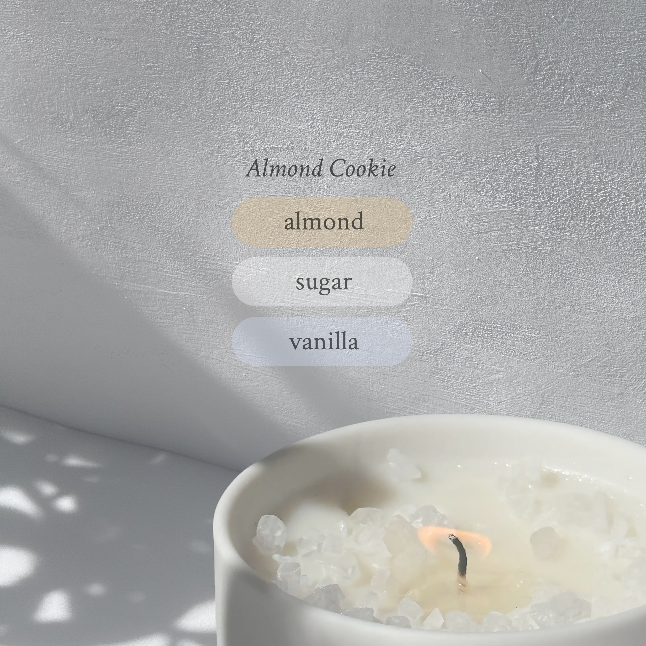 Large stoneware candle, lit, it’s top covered in rock salt. The title reads “Almond Cookie”, and underneath is the scent profile: almond, sugar, vanilla.