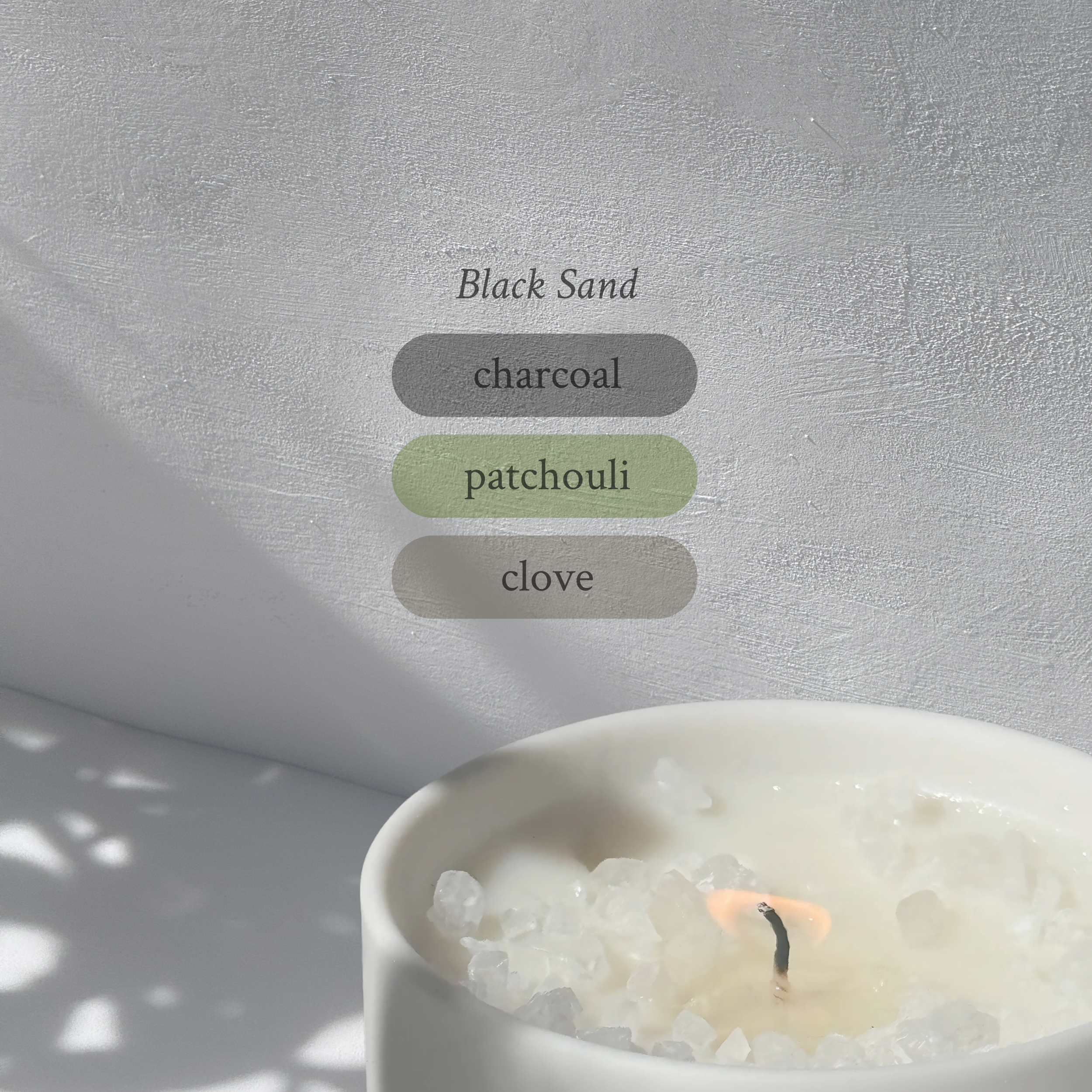 Large stoneware candle, lit, it’s top covered in rock salt. The title reads “Black Sand”, and underneath is the scent profile: charcoal, patchouli, clove.