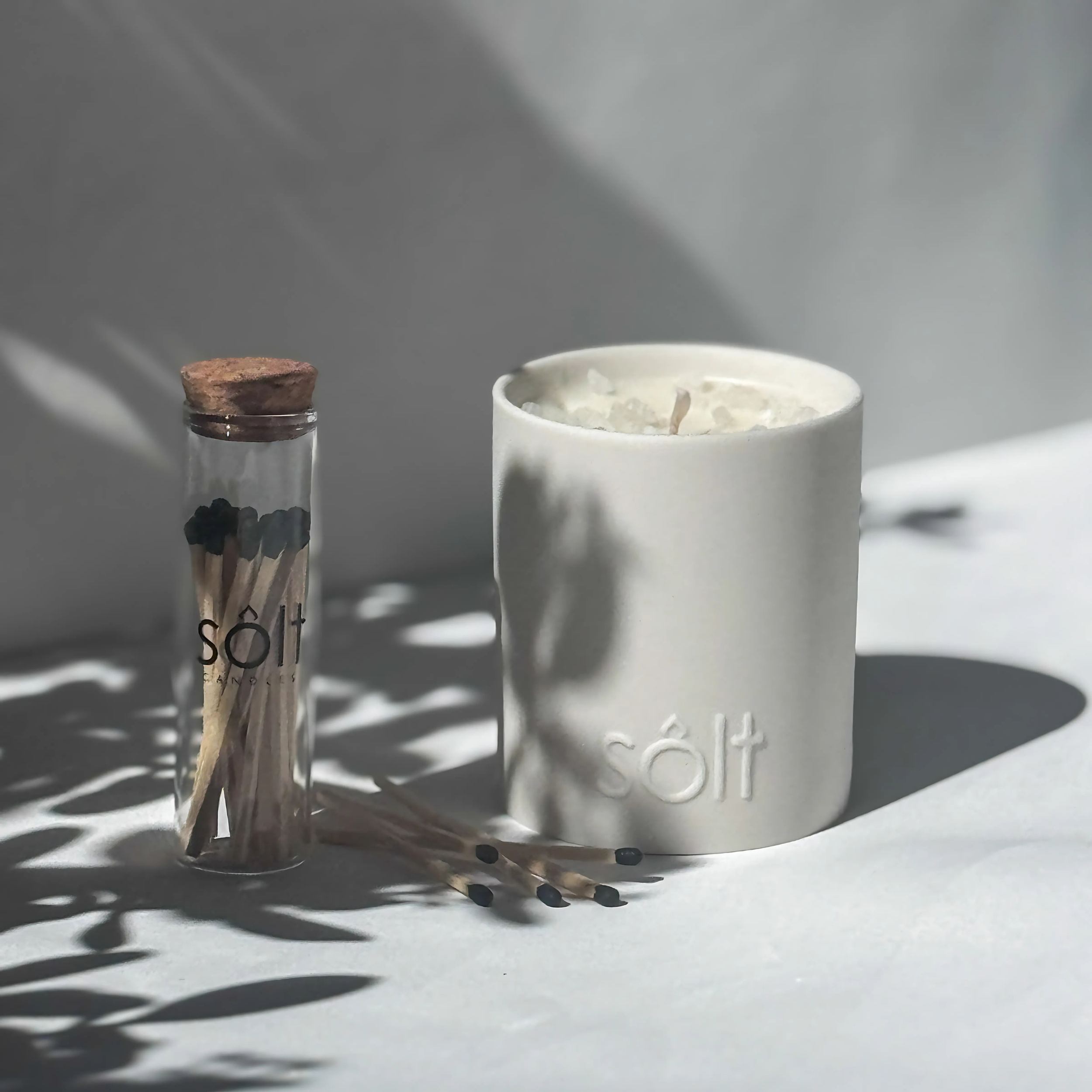A large stoneware candle sits unlit next to a match jar. A few loose matches sprinkle the ground.