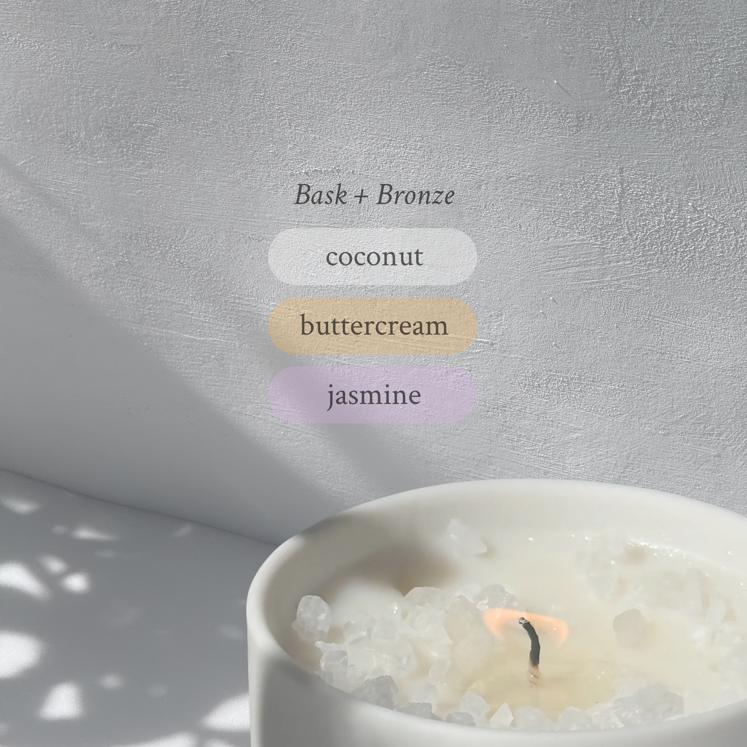 Large stoneware candle, lit, it’s top covered in rock salt. The title reads “Bask + Bronze”, and underneath is the scent profile: coconut, buttercream, jasmine.