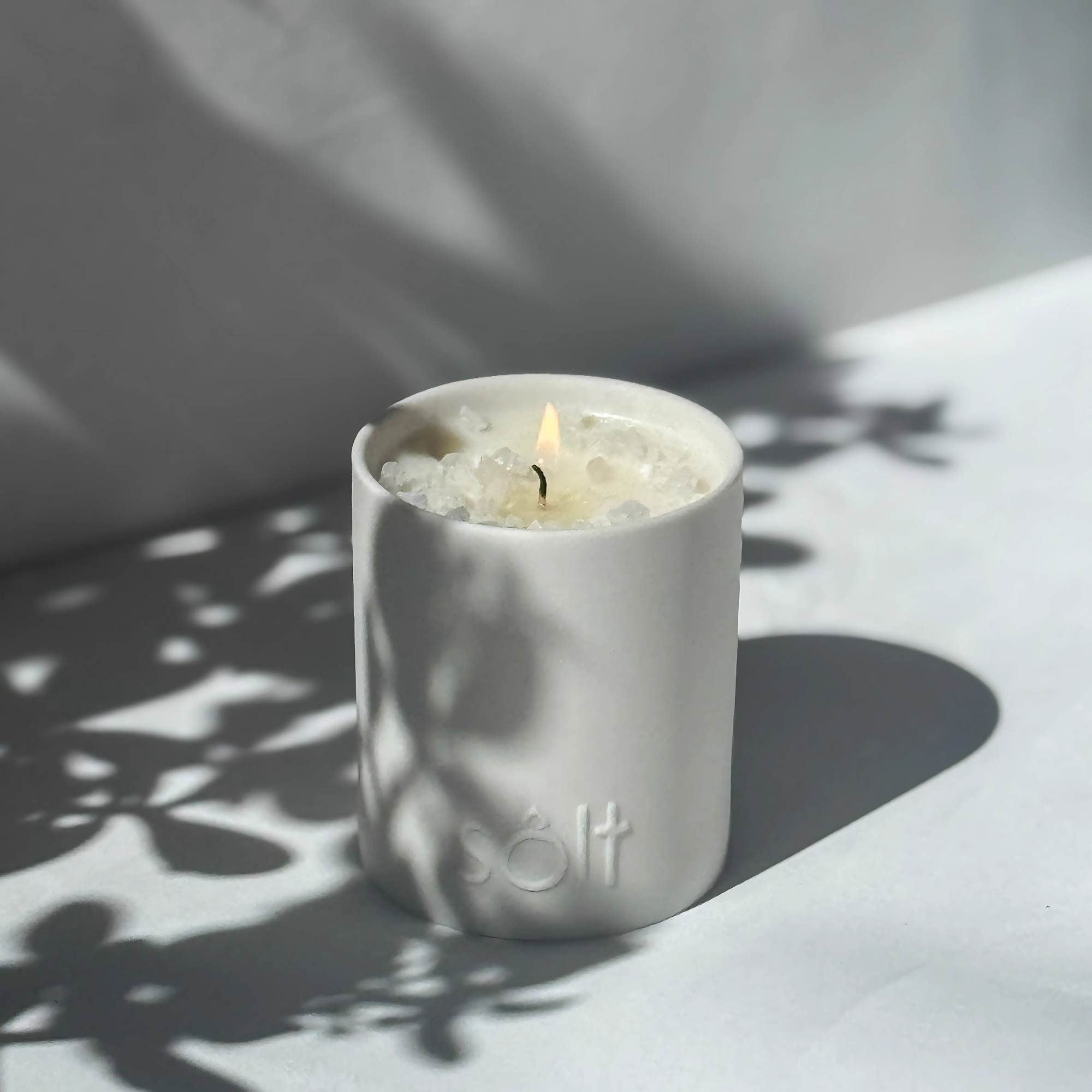 Large stoneware candle, lit in a sun dappled area. The top of the candle is crystallized with rock salt, and a pool of wax grows.