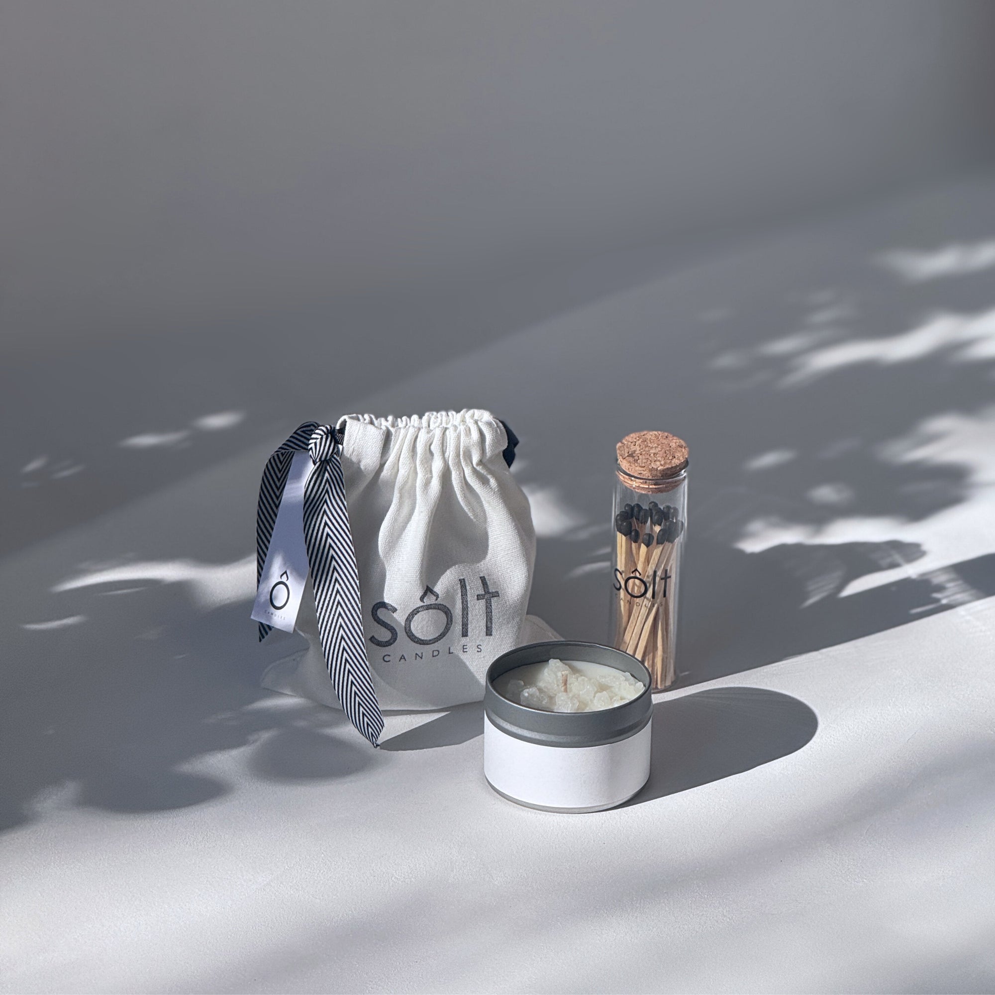 Image of a SOLT gift set, including a mini candle tin covered in crystallized rock salt, a glass jar of matches, and a soft canvas gift bag tied with a ribbon.