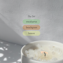 Large stoneware candle, lit, it’s top covered in rock salt. The title reads “The Del”, and underneath is the scent profile: eucalyptus, beachgrass, lemon.