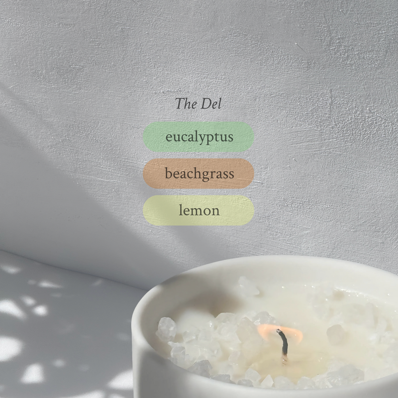 Large stoneware candle, lit, it’s top covered in rock salt. The title reads “The Del”, and underneath is the scent profile: eucalyptus, beachgrass, lemon.